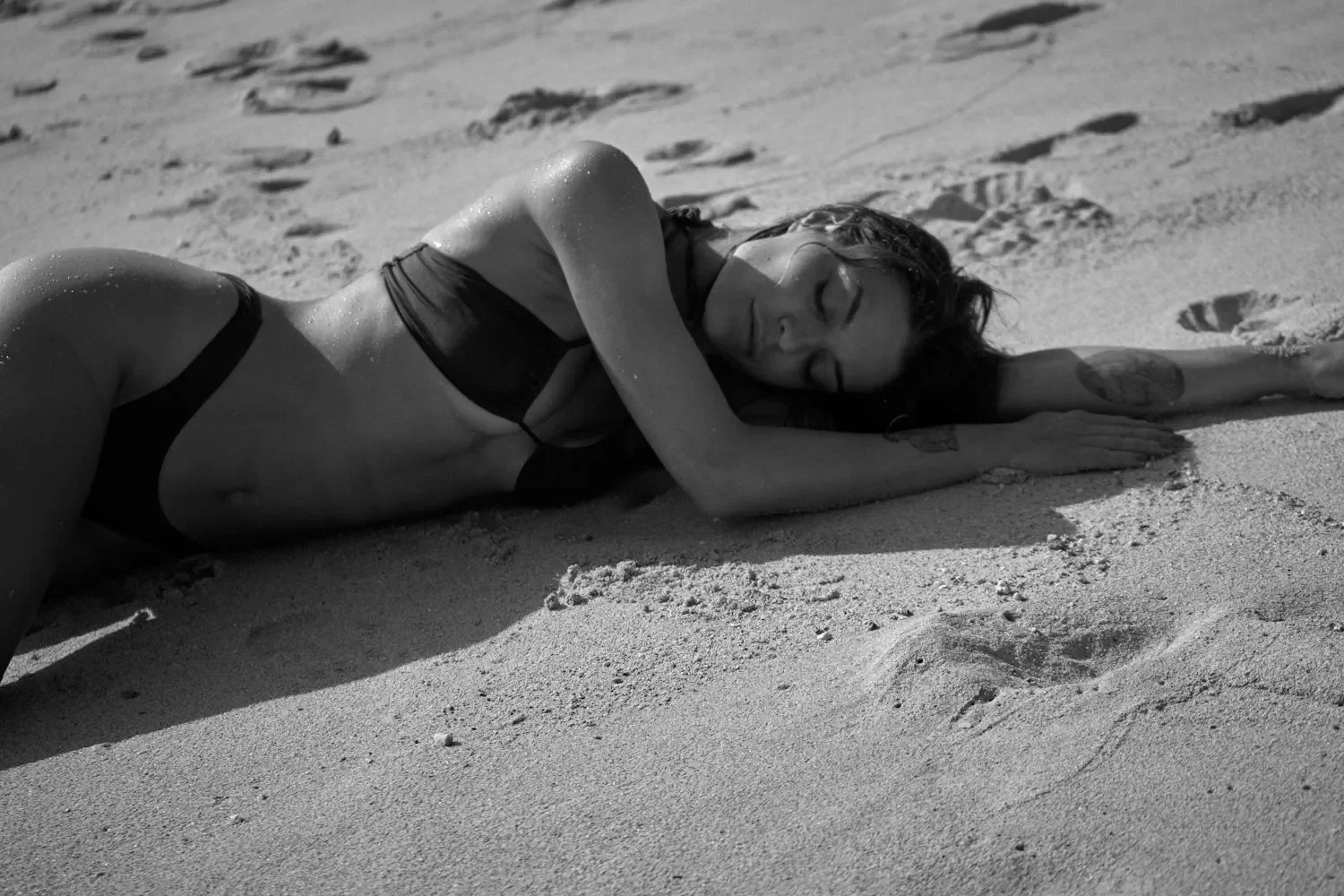 Mujer descansando en la playa, acostada sobre la arena con un bikini negro, en una escena en blanco y negro que transmite tranquilidad y relajación.